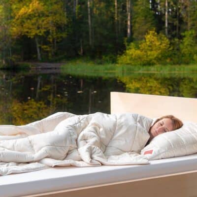 Eine Person schläft friedlich auf einer Sommerdecke aus reiner Wolle von HEFEL im Freien an einem ruhigen See, eingehüllt in weiße Bettwäsche und umgeben von grünen und gelben Bäumen.