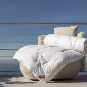 Eine kuschelige weiße Bettdecke und ein passendes Kissen sind über einem beigefarbenen Sessel auf einem Balkon drapiert, mit einem Metallgeländer und einem klaren blauen Himmel im Hintergrund.