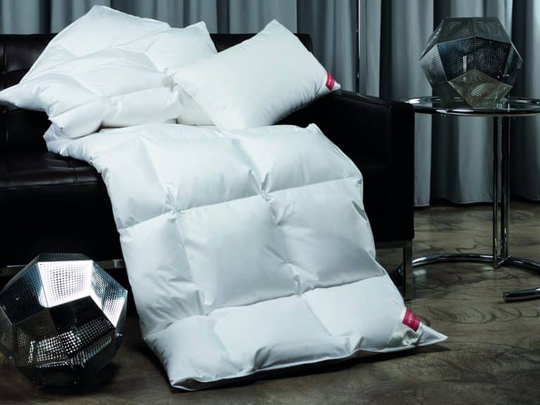 Weiße Kissen und eine bauschige weiße Bettdecke liegen auf einem dunklen Sofa und sind auf dem Holzboden drapiert; daneben stehen zwei geometrische Metalldekorationen und ein runder Beistelltisch aus Glas, mit grauen Vorhängen im Hintergrund.