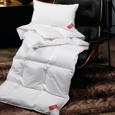 Die Daune Gold Down Ganzjahresdecke von HEFEL mit roten Anhängern und einem weißen Kissen ist über einen modernen schwarzen Sessel auf einem braunen Fellteppich drapiert, in einem stilvollen Raum mit einem dunklen Schrank im Hintergrund.