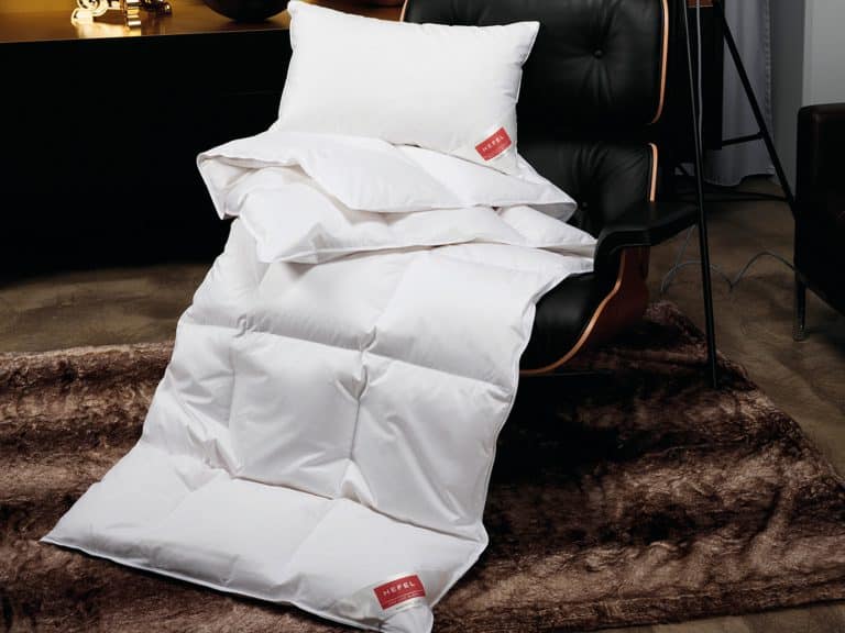 Die Daune Gold Down Ganzjahresdecke von HEFEL mit roten Anhängern und einem weißen Kissen ist über einen modernen schwarzen Sessel auf einem braunen Fellteppich drapiert, in einem stilvollen Raum mit einem dunklen Schrank im Hintergrund.