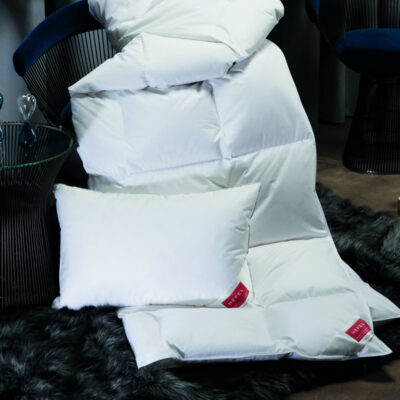 Eine weiße Bettdecke und Kissen sind auf einem grauen Fellteppich neben einem gläsernen Beistelltisch mit Sanduhren und blauen Samtstühlen in einem modernen, gemütlichen Raum angeordnet.