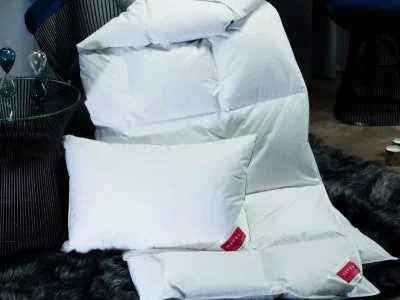 Ein weißes Kissen und die KlimaControl Comfort Winterdecke von HEFEL liegen auf einem dunklen Kunstfellteppich neben einem modernen Glasbeistelltisch, im Hintergrund sind Sanduhren und Stühle zu sehen.