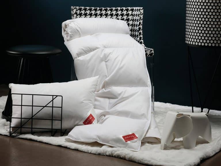 Eine gemütliche Szene mit der Daune DeLuxe Daunen-Ganzjahresdecke von HEFEL, einer weißen Bettdecke, einem Kissen und einem Kissen mit roten Anhängern auf einem flauschigen Teppich sowie einem schwarzen Hocker, einem Stuhl mit Hahnentrittmuster, einem Drahtkorb und einem Dekor in Elefantenform.