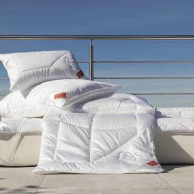 A neatly made bed with white pillows and a duvet, stacked and arranged on a terrace with metal railings, against a clear blue sky.