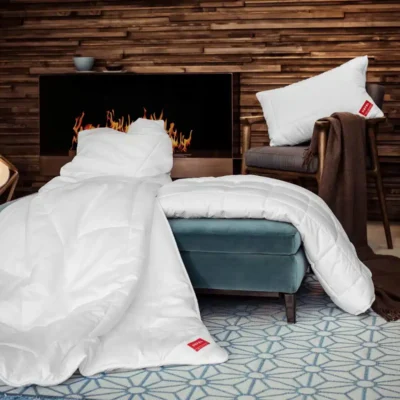 Ein gemütliches Wohnzimmer mit einem modernen Kamin, einem blauen Hocker mit der KlimaControl Comfort Winterdecke von HEFEL, einem weißen Kissen auf einem braunen Stuhl und einer weichen Decke auf einem geometrisch gemusterten Teppich.