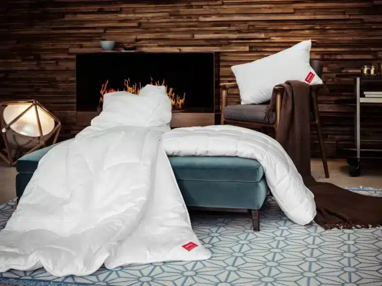 Ein gemütliches Wohnzimmer mit einem modernen Kamin, einem blauen Hocker mit der KlimaControl Comfort Winterdecke von HEFEL, einem weißen Kissen auf einem braunen Stuhl und einer weichen Decke auf einem geometrisch gemusterten Teppich.