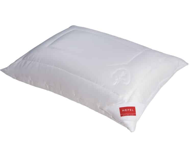 Ein weißes, rechteckiges Kissen mit einem dezenten, abgesteppten Baummuster in der Ecke und einem roten Etikett mit der Aufschrift HEFEL unten rechts. Das Kissen hat eine glatte, gesteppte Oberfläche.