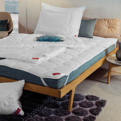 A neatly made bed with a white quilted duvet, matching pillows and a blue accent pillow. The wooden bed frame stands next to a small nightstand with a vase and candles, and a patterned rug lies on the floor.