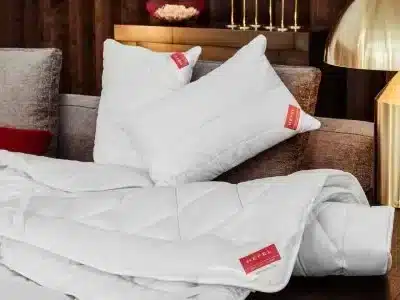 Weiße Kissen und eine gefaltete KlimaControl Comfort Winterdecke von HEFEL mit rotem Etikett sind auf einem beigen Sofa angeordnet, während eine goldene Lampe und dekorative Akzente im warm beleuchteten Hintergrund erscheinen.