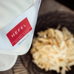 Eine Nahaufnahme einer weißen Stoffecke mit rotem "Hefel Bed and Sleep"-Etikett über einem unscharfen Korb aus leicht zerkleinertem Material zeigt die Bio Zirbe Ganzjahresdecke von HEFEL.