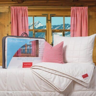 Ein gemütliches Schlafzimmer mit holzgetäfelten Wänden, roten Gingham-Vorhängen und einem Fenster, das verschneite Berge zeigt. Auf dem Bett liegen eine weiße Bettdecke, ein Kissen und ein dazu passendes kariertes Kissen. Die verpackte Bettwäsche ist mit HEFEL beschriftet.