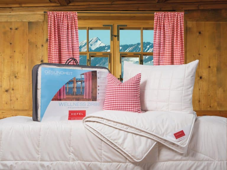A cozy bedroom with wood-panelled walls, red gingham curtains and a window showing snow-covered mountains. On the bed are a white comforter, a pillow and a matching plaid pillow. The wrapped bed linen is labeled HEFEL.