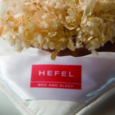 Eine Hand hält eine Mischung aus weichem weißem und hellbraunem Füllmaterial über einer weißen Bettdecke mit einem roten Etikett mit der Aufschrift HEFEL BED AND SLEEP.