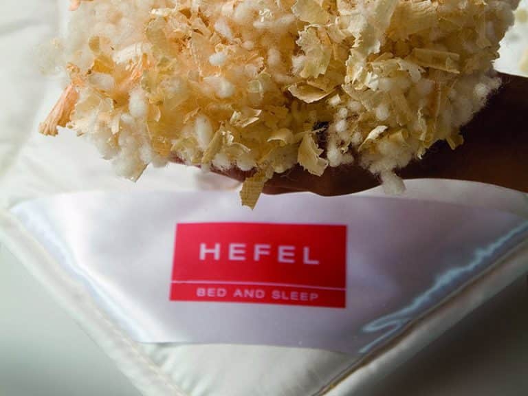 One hand holds a mixture of soft white and light brown filling material over a white comforter with a red label bearing the words HEFEL BED AND SLEEP.