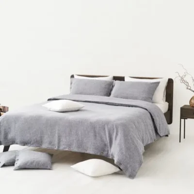 A minimalist bedroom with a bed dressed in Cygna Classic Linen Sheet by Geniksa, light grey pillows, a wooden nightstand with a vase of branches, additional pillows and a woven basket on the white floor.