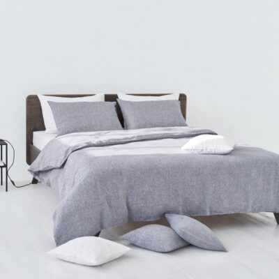 In a minimalist room, a neatly made bed with Heron Classic Linen Bedding by Geniksa in grey is complemented by white pillows. Next to the bed is a nightstand with a lamp and a book, and pillows are scattered on the white wooden floor.