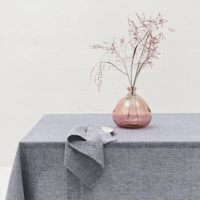 A pink glass vase with dried branches on a table set with the Heron linen tablecloth by Geniksa. A matching grey napkin is draped over the edge against a plain white wall.