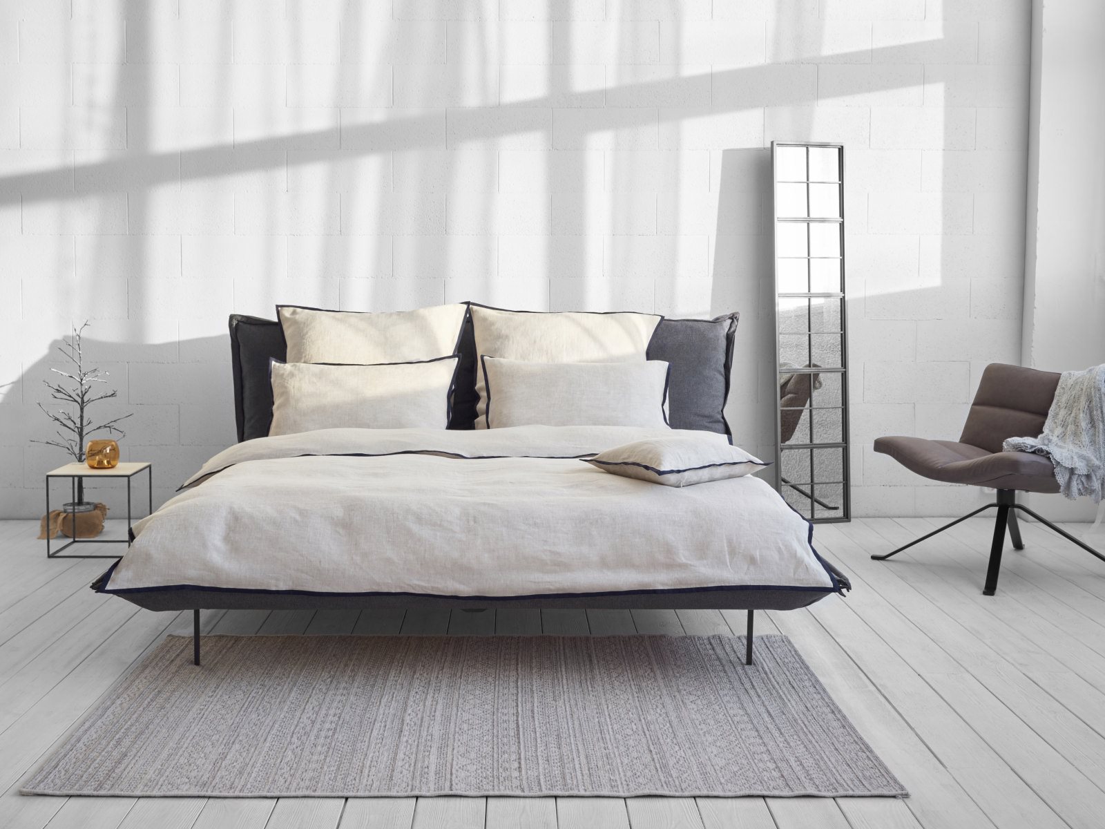 Ein modernes, minimalistisches Schlafzimmer mit einem sauber gemachten Bett aus Hirundo Leinen Bettwäsche von Geniksa, neutraler Bettwäsche, einem Nachttisch mit einer Pflanze, einem Bodenspiegel, einem braunen, mit einer Decke drapierten Stuhl und Sonnenlicht, das durch das Fenster fällt.