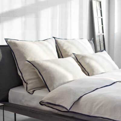 A modern, sun-filled bedroom with a neatly made bed featuring Hirundo linen bedding by Geniksa in cream and navy blue, four large pillows, a duvet and a mirror on the wall.