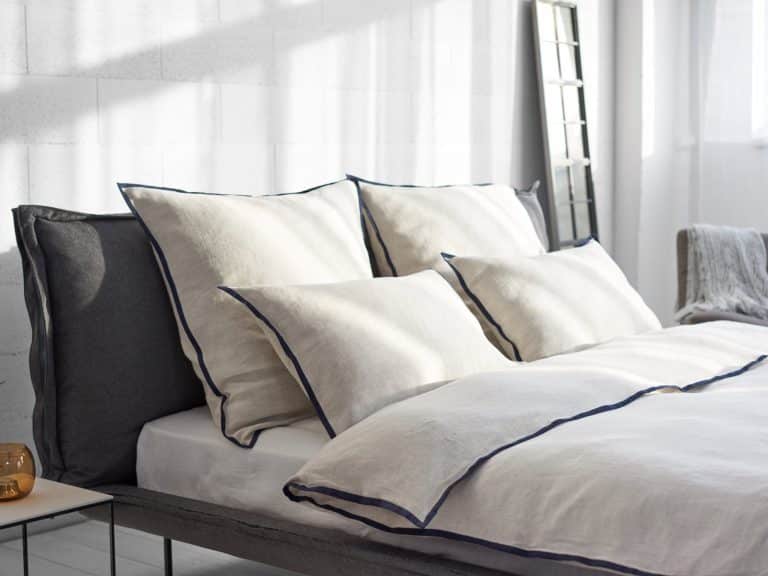 A modern, sun-filled bedroom with a neatly made bed featuring Hirundo linen bedding by Geniksa in cream and navy blue, four large pillows, a duvet and a mirror on the wall.