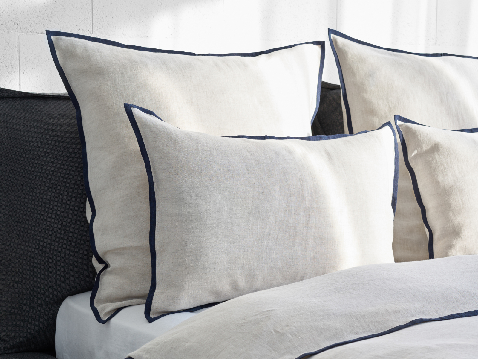Drei beigefarbene Leinenkissen mit dunkelblauen Paspeln liegen auf einem mit Hirundo Leinen Bettwäsche von Geniksa bezogenen Bett, in das das Sonnenlicht einfällt und das eine helle und gemütliche Atmosphäre schafft.