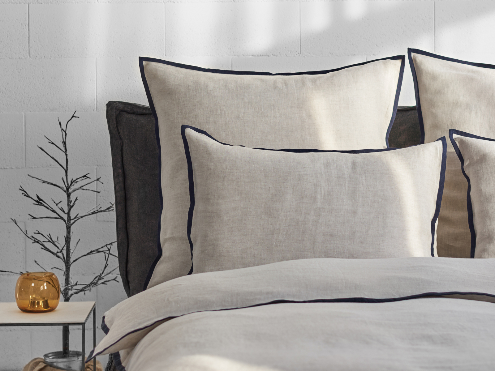 Ein ordentlich gemachtes Bett mit beiger Hirundo Leinen Bettwäsche von Geniksa mit dunkler Paspelierung steht vor einer hellen Wand. Daneben steht ein kleiner Tisch mit einem gläsernen Kerzenständer und einem minimalistischen Zweigdekor, wobei das Sonnenlicht weiche Schatten an die Wand wirft.