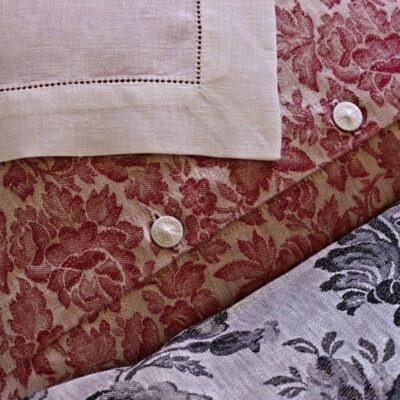 Eine Nahaufnahme von Chrysantheme Rot Leinen Damast von Hoffmann zeigt rote und graue Blumenmuster, zwei weiße Knöpfe und ein Stück beiges Leinen mit Ziernähten in der linken oberen Ecke.