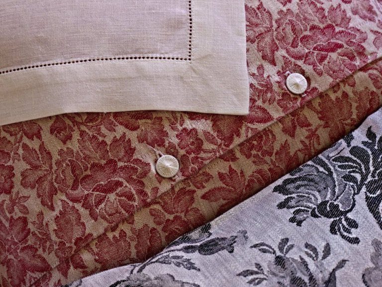 Eine Nahaufnahme von Chrysantheme Rot Leinen Damast von Hoffmann zeigt rote und graue Blumenmuster, zwei weiße Knöpfe und ein Stück beiges Leinen mit Ziernähten in der linken oberen Ecke.