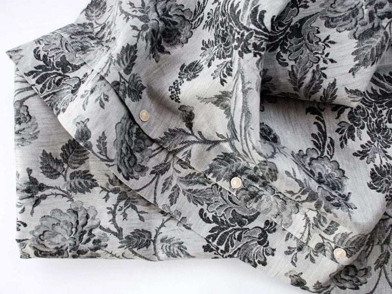 Ein hellgraues, gefaltetes Hemd aus Chrysantheme Anthrazit Leinen Damast von Hoffmann, das ein strukturiertes schwarzes Blumenmuster aus Blättern und Rosen zeigt und mit weißen Knöpfen versehen ist.