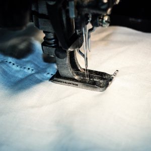 Nahaufnahme einer Nähmaschinennadel, die einen weißen Stoff vernäht, mit detaillierten Fäden und Stoffstruktur beim Niederdrücken des Fußes.