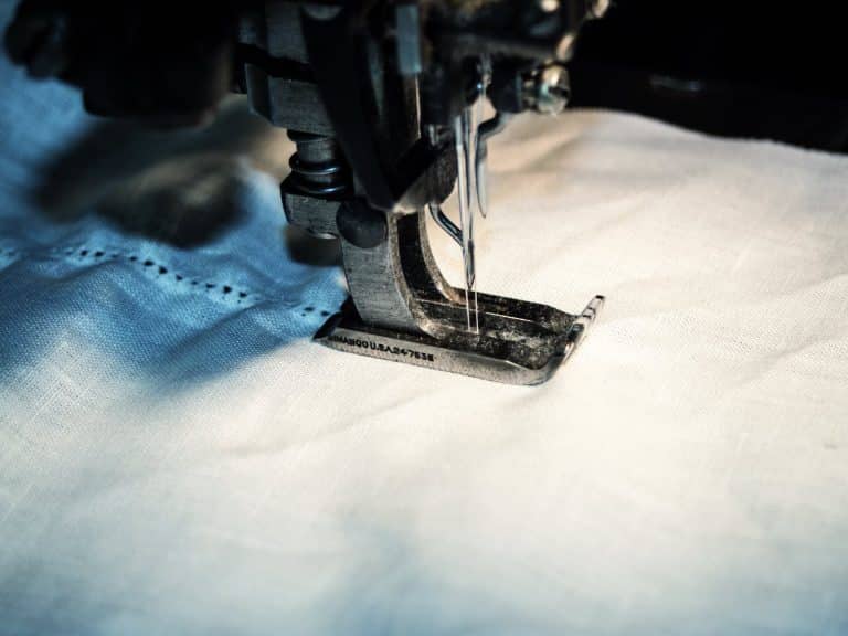 Nahaufnahme einer Nähmaschinennadel, die einen weißen Stoff vernäht, mit detaillierten Fäden und Stoffstruktur beim Niederdrücken des Fußes.