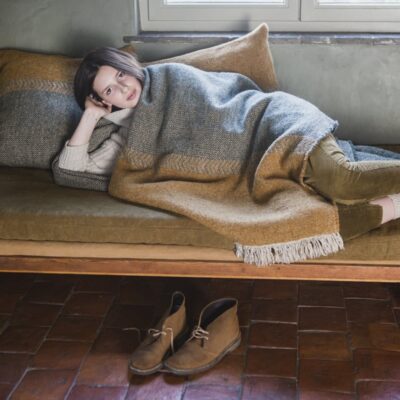 Eine Person entspannt sich auf einem mit der Decke Jules Green Herringbone Leinen-Wolle-Mix von Libeco drapierten Sofa, den Kopf auf einem Kissen, und schaut in die Kamera, während ihre Schuhe ordentlich auf dem gefliesten Boden stehen.
