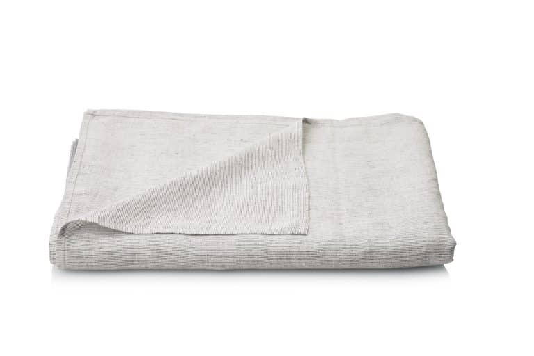 A neatly folded light gray Junco linen bed sheet from Geniksa with a subtle textured pattern is shown on a white background with one corner turned up slightly to show the underside.