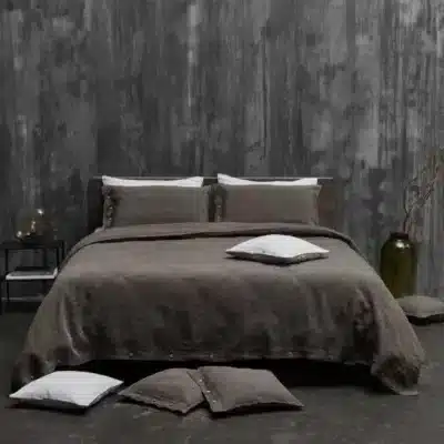 A neatly made bed against a textured grey wall is dressed with luxurious Cygna Classic Linen Sheet by Geniksa, dark brown bedding and matching pillows. On a small side table are a lamp, a glass, and nearby a vase with branches.