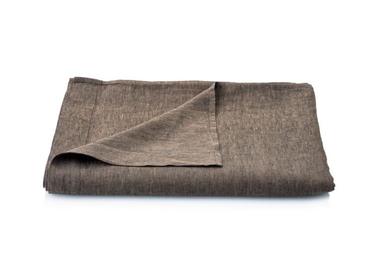 A folded brown cloth napkin with a raised corner is depicted on a white background and features earth tones inspired by Geniksa's jungle linen bed sheets.