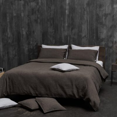 In a modern bedroom, a bed with dark brown Jungle Classic Linen Bedding by Geniksa stands against a textured grey wall. Several pillows lie on the floor, and next to the bed is a small nightstand with a vase.