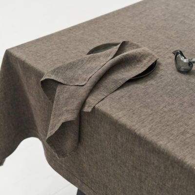 Close-up of a table with the Jungle linen tablecloth by Geniksa, a matching folded napkin on a dark plate, a small glass bird figurine and stacked brown rectangular objects in the corner.