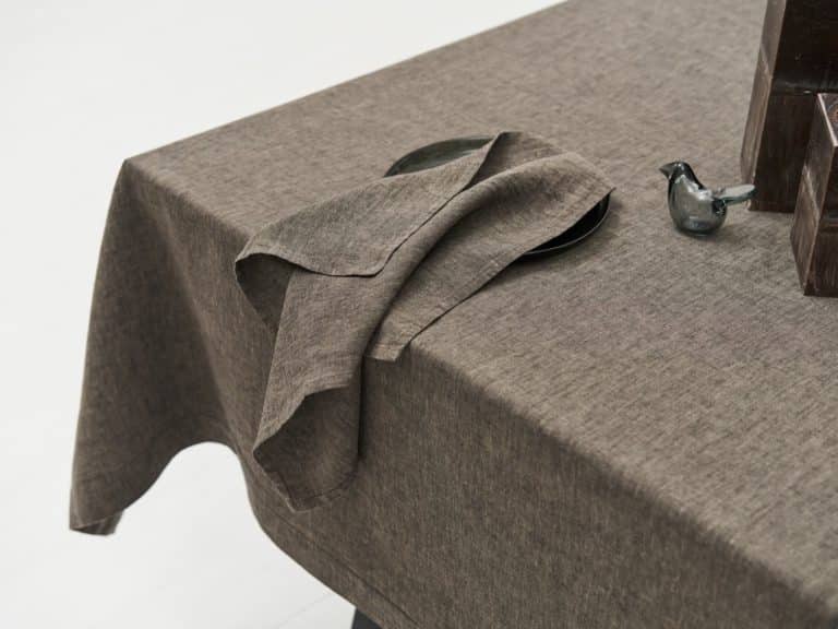 Close-up of a table with the Jungle linen tablecloth by Geniksa, a matching folded napkin on a dark plate, a small glass bird figurine and stacked brown rectangular objects in the corner.