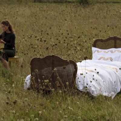 Ein Vintage-Bett, bezogen mit Laurels Percal 400TC Bettwäsche von Peter Reed in Weiß und Gold, steht in einem mit Wildblumen übersäten Feld, während eine Frau in der Nähe auf einem Stuhl sitzt und liest.