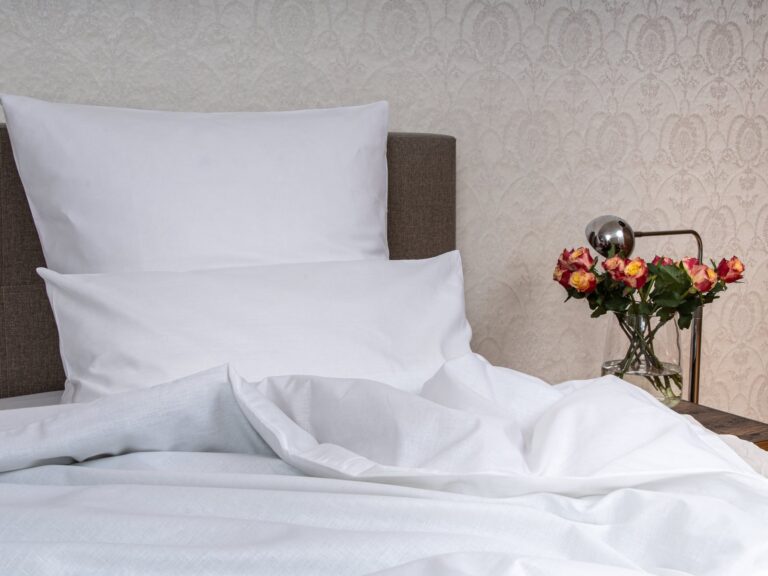 A bed with white pillows and white half-linen bedding from the Hoffmann linen weaving mill stands next to a wooden bedside table with a glass vase of pink and yellow roses in front of a patterned cream-colored wallpaper.