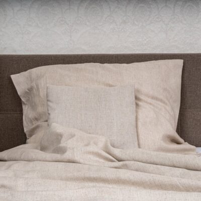 Ein Bett mit beiger Natur Leinen Bettwäsche der Leinenweberei Hoffmann - mit einem großen Kissen, einem quadratischen Kissen und einer Bettdecke - steht vor einem braun gepolsterten Kopfteil und einer weißen Strukturtapete, mit einer Pflanze zur Linken.