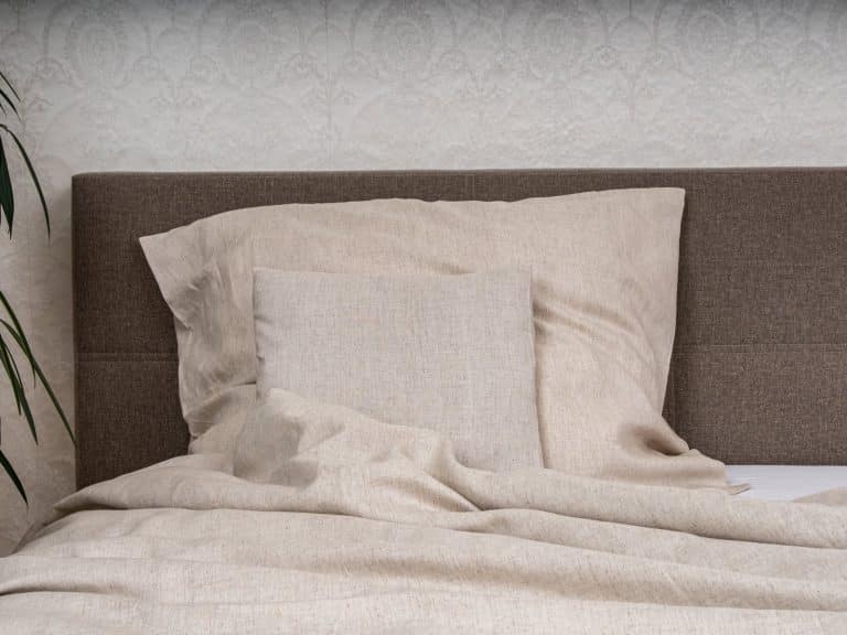 A bed with beige natural linen bedding from Leinenweberei Hoffmann - with a large cushion, a square cushion and a comforter - stands in front of a brown upholstered headboard and white textured wallpaper, with a plant to the left.
