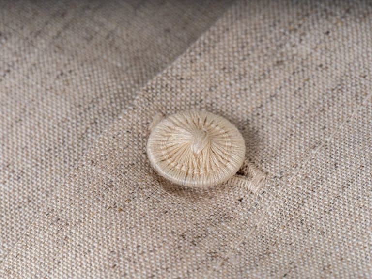 A close-up shows a beige fabric-covered button sewn onto a matching textured fabric with a fine seam - an ideal complement to the natural linen bed linen from Leinenweberei Hoffmann.