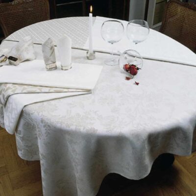 Ein runder Tisch mit der Tischdecke aus feinem weißen Leinen von der Leinenweberei Hoffmann, gedeckt mit zwei leeren Weingläsern, einer brennenden Kerze, gefalteten Servietten, einem Glas mit einer roten Rose und umgeben von zwei Holzstühlen.