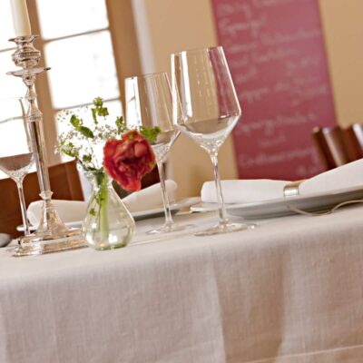 Ein eleganter Restauranttisch ist mit Weingläsern, Tellern, weißen Servietten und Blumen auf einer Tischdecke aus feinem weißen Leinen von der Leinenweberei Hoffmann gedeckt. Im Hintergrund erscheinen Stühle und ein Menübrett sanft verschwommen.