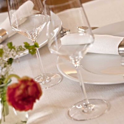 Ein stilvoller Tisch mit weißen Tellern, silbernen Serviettenringen mit weißen Servietten, Weingläsern und einem kleinen Blumenarrangement auf einer Tischdecke aus feinem weißen Leinen von der Leinenweberei Hoffmann.