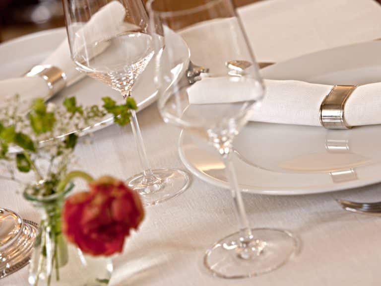A stylish table with white plates, silver napkin rings with white napkins, wine glasses and a small flower arrangement on a tablecloth made of fine white linen from the Hoffmann linen weaving mill.