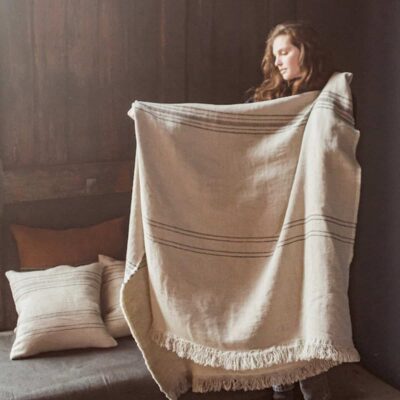 Eine Frau im Haus hält eine große, fransenbesetzte Marrakesch Leinen-Wolle-Mix von Libeco-Decke mit dunklen Streifen hoch. Hinter ihr liegen passende Kissen auf einem Bett und einer Holzwand, während von links Tageslicht einfällt.