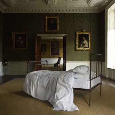 Ein ordentlich gemachtes Bett mit der Matisse Percal 400TC Bettwäsche von Peter Reed steht in einem großen, schwach beleuchteten Raum mit verzierten Wänden und Porträts in Goldrahmen, die ein elegantes, historisches Ambiente schaffen.